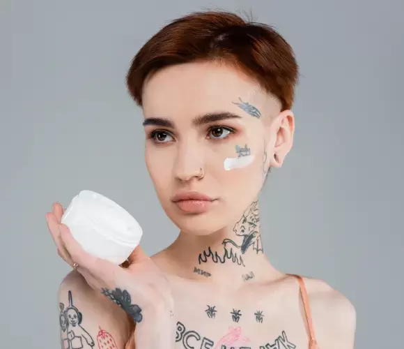 Person with tattoos holding skincare product, showing aftercare application
