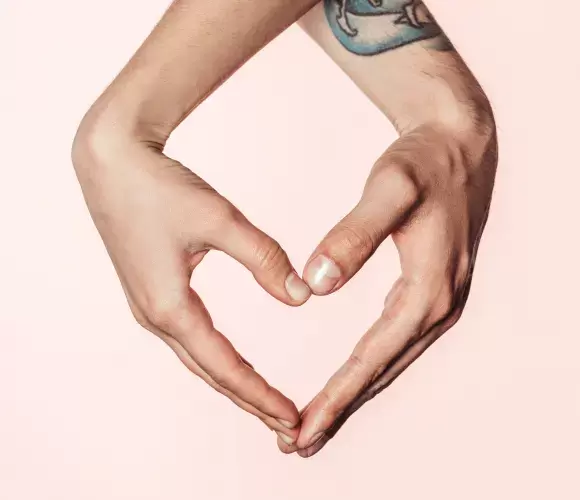 Two hands forming a heart shape on pale pink background with small wrist tattoo